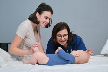 Pediatrician woman doctor in blue uniform with stethoscope talking to young mother of little boy, examining 7 month old baby at home