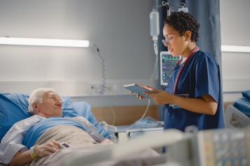 Friendly Female Head Nurse Making Rounds does Checkup on Elderly Patient Resting in Bed.  She Checks Computer for Vitals while Old Man Fully Recovering after Successful Surgery.