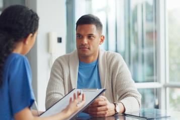 A female doctor with long hair in a tail talking to a young male patient in a blue t-shirt and beige cardigan in a hospital setting.