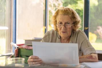 Older woman wearing eyeglasses, reading documents while sitting at a table at home.