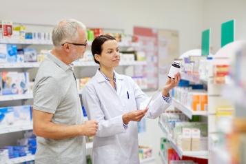 A pharmacist in a white coat stands beside an older man in a pharmacy aisle, holding up a bottle of medication and explaining the label while checking a prescription sheet. Shelves of medicines and health products line the background.