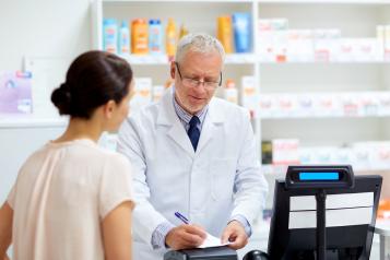 A pharmacist in a white coat stands behind a pharmacy counter, writing on a prescription form while speaking with a woman on the other side. Shelves of medicines and health products are visible in the background.
