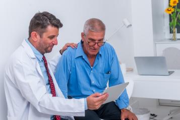 Doctor in a white coat sits beside an older man in a blue shirt, resting a reassuring hand on his shoulder while showing him information on a tablet. They are in a bright clinical room with a desk, laptop, and sunflowers in the background.