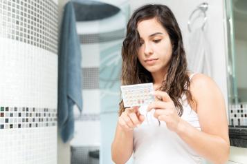 A woman standing in a bathroom looks down at a packet of contraceptive pills in her hands. She is wearing a white top, with tiled walls and a towel hanging in the background.