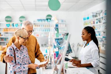 An older couple stand at the counter of a bright community pharmacy, speaking with a pharmacist in a white uniform. The woman holds a prescription slip while the man stands beside her with his arm around her shoulder. Shelves of medicines and health products are visible in the background.