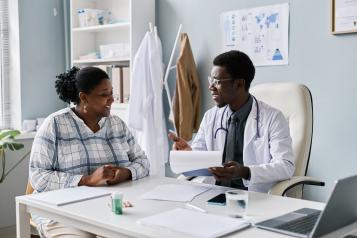 Doctor sitting at a desk in a GP surgery smiles and speaks with a woman during a consultation, holding paperwork. The room contains medical posters, shelves, a stethoscope around the doctor’s neck, and a laptop on the desk.
