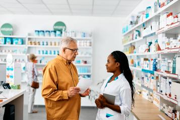 An older man holding a prescription sheet speaks with a pharmacist in a white coat inside a bright community pharmacy. Shelves of medicines and health products line the walls, and another customer is visible in the background browsing items.