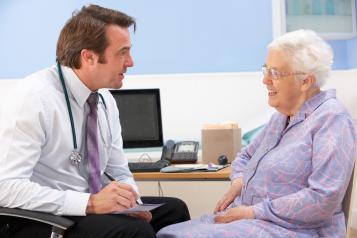 Doctor talking to an older female patient in doctor's surgery. He is writing a prescription and she is smiling whilst looking at him.