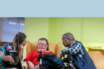 Three people sitting together in a care setting having a conversation. An older woman in a red jumper is speaking with two people beside her, with a walking frame visible in the foreground.