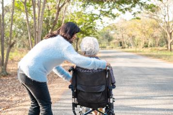 An unpaid carer pushing an older person in a wheelchair along a peaceful tree-lined path.