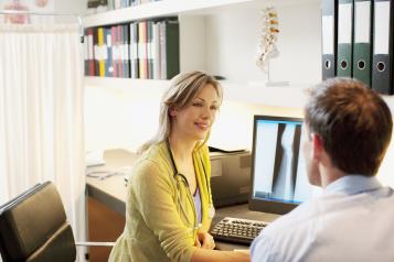 Female healthcare professional wearing a stethoscope talking with a male patient in a GP consultation room.
