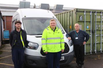 Three members of the hospital transport team stand beside a white minibus outside a hospital service building. The person in the centre is wearing a bright yellow high-visibility jacket, while the two others wear dark work clothing. Large green storage containers and hospital buildings can be seen behind them.
