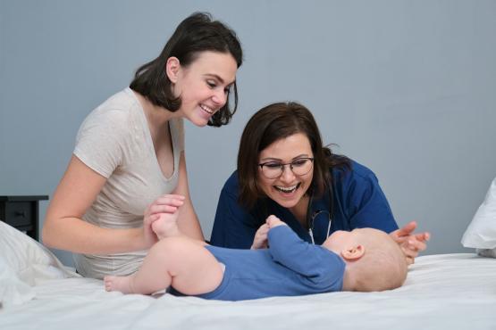 Pediatrician woman doctor in blue uniform with stethoscope talking to young mother of little boy, examining 7 month old baby at home