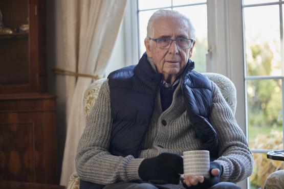 Older man wearing extra clothes with hot drink trying to keep warm at home in an energy crisis
