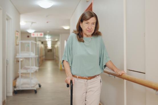 Older woman with a disability holding a railing on a hospital corridor whilst waiting for care.