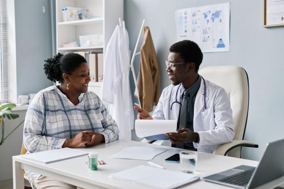 Doctor sitting at a desk in a GP surgery smiles and speaks with a woman during a consultation, holding paperwork. The room contains medical posters, shelves, a stethoscope around the doctor’s neck, and a laptop on the desk.