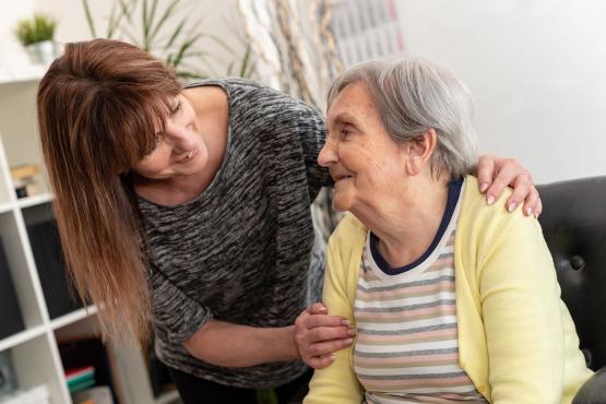 A woman places a reassuring hand on the shoulder of an older woman as they smile and make eye contact while talking in a comfortable home setting. The image represents care, support and companionship for older people.