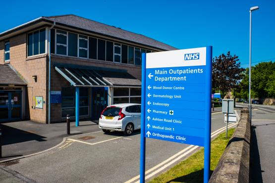 Exterior of an NHS hospital outpatient department on a bright sunny day. A large blue and white NHS direction sign stands in the foreground, pointing towards the Main Outpatients Department, Blood Donor Centre, Dermatology Unit, Endoscopy, Pharmacy, Ashton Road Clinic, Medical Unit 1 and Orthopaedic Clinic. The hospital building and entrance are visible behind the sign, with a car parked outside and a road running alongside the site.