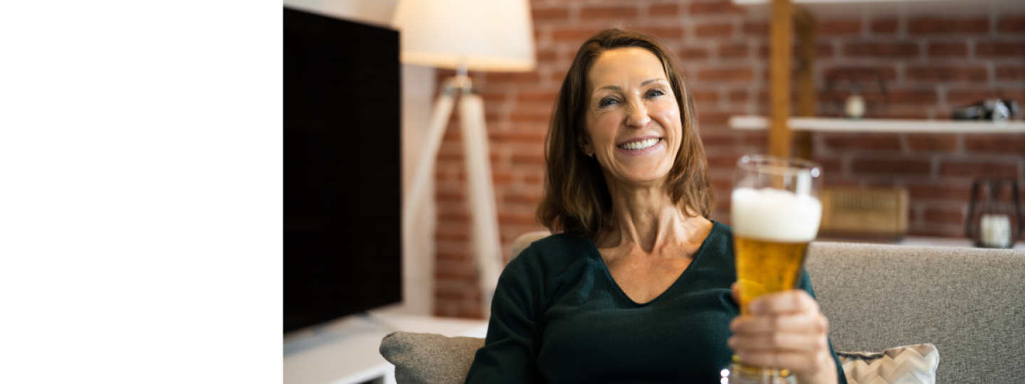 A midlife lady smiling whilst sitting on a couch holding up a glass of beer