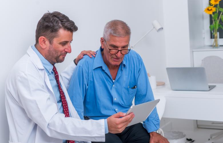 Doctor in a white coat sits beside an older man in a blue shirt, resting a reassuring hand on his shoulder while showing him information on a tablet. They are in a bright clinical room with a desk, laptop, and sunflowers in the background.