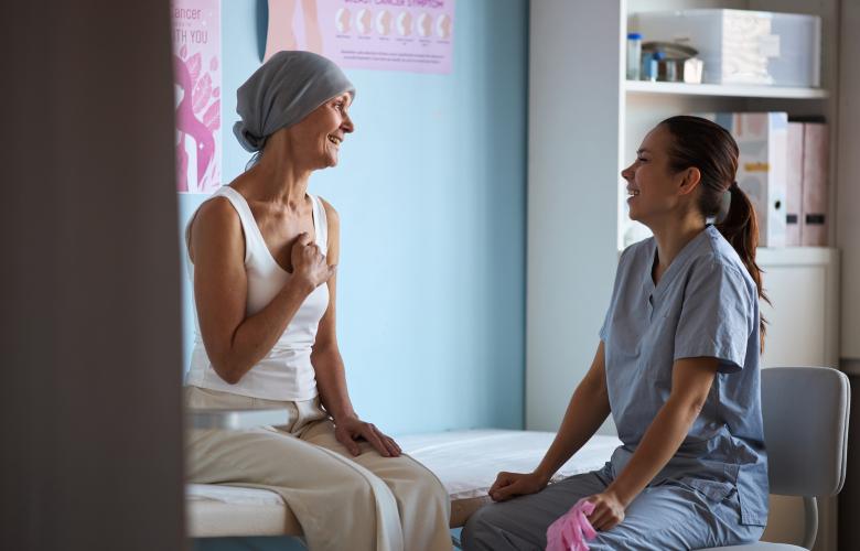 Middle aged woman with headscarf sitting on examination table, smiling and talking with young adult female nurse, in medical clinic with breast cancer awareness posters visible