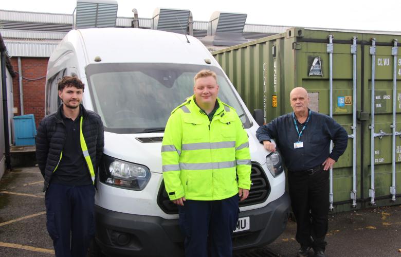Three members of the hospital transport team stand beside a white minibus outside a hospital service building. The person in the centre is wearing a bright yellow high-visibility jacket, while the two others wear dark work clothing. Large green storage containers and hospital buildings can be seen behind them.