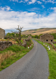 A rural road in Hawes, North Yorkshire - with fields at either side and two stone outbuildings.