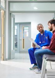 A male doctor sat in a hospital corridor talking with a male patient in his twenties.