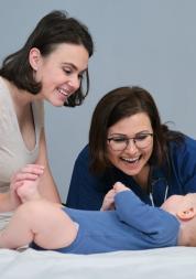Pediatrician woman doctor in blue uniform with stethoscope talking to young mother of little boy, examining 7 month old baby at home