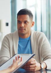 A female doctor with long hair in a tail talking to a young male patient in a blue t-shirt and beige cardigan in a hospital setting.