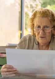 Older woman wearing eyeglasses, reading documents while sitting at a table at home.