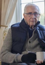 Older man wearing extra clothes with hot drink trying to keep warm at home in an energy crisis