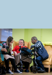Three people sitting together in a care setting having a conversation. An older woman in a red jumper is speaking with two people beside her, with a walking frame visible in the foreground.